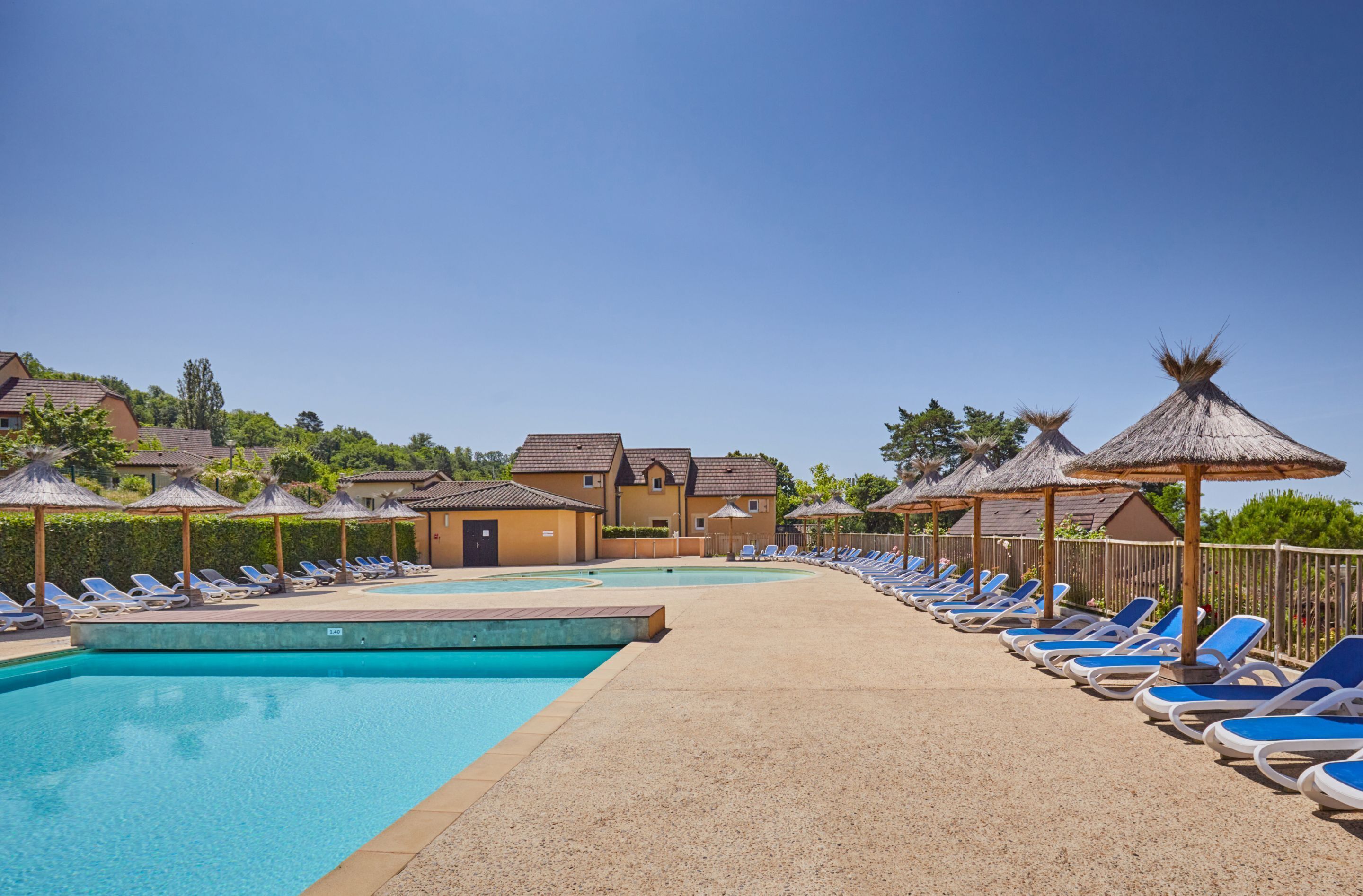 A pool area with lounge chairs and umbrellas, surrounded by buildings with thatched roofs.