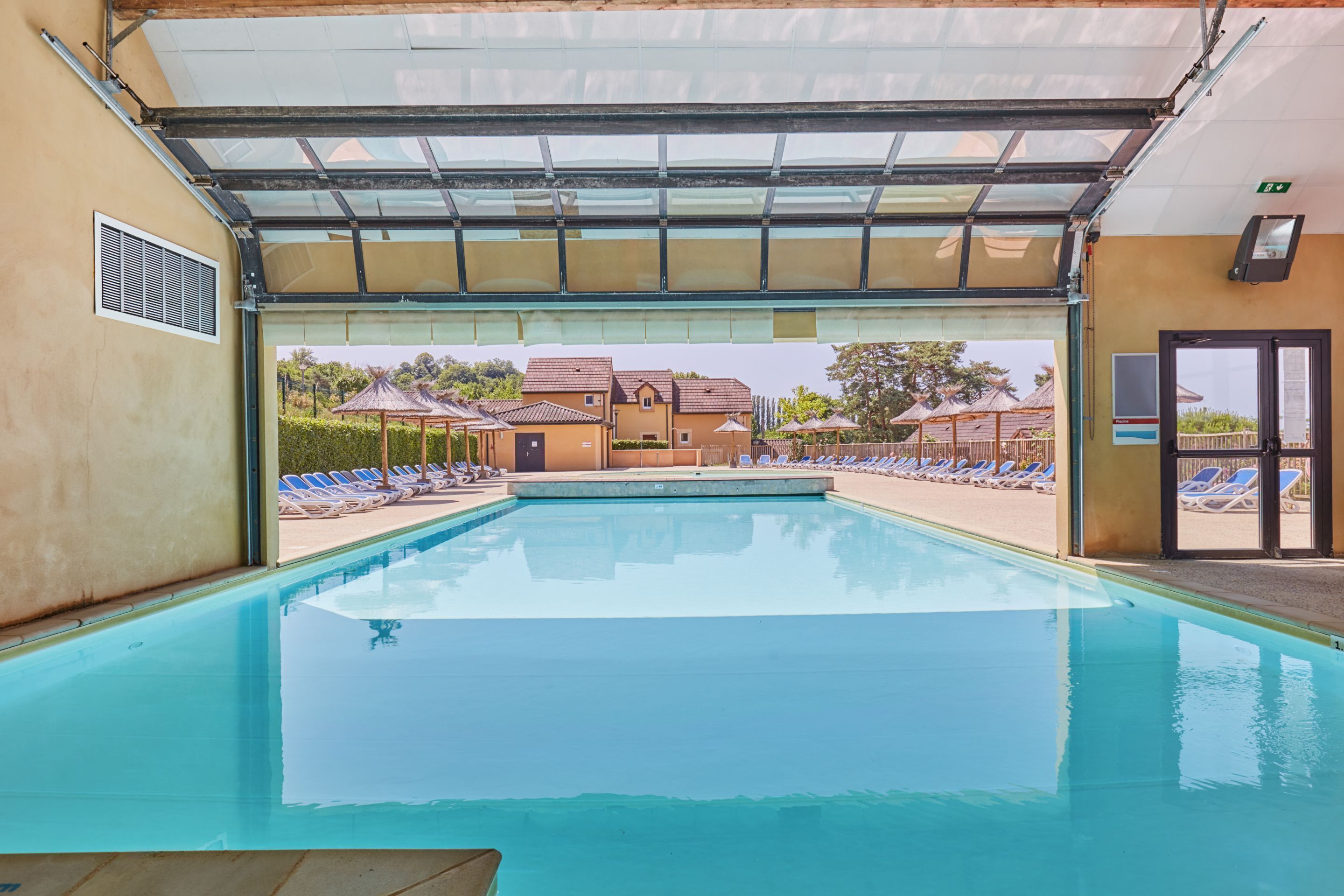 Una piscina interior con paredes de vidrio transparente, vistas a una zona residencial y una entrada cubierta.