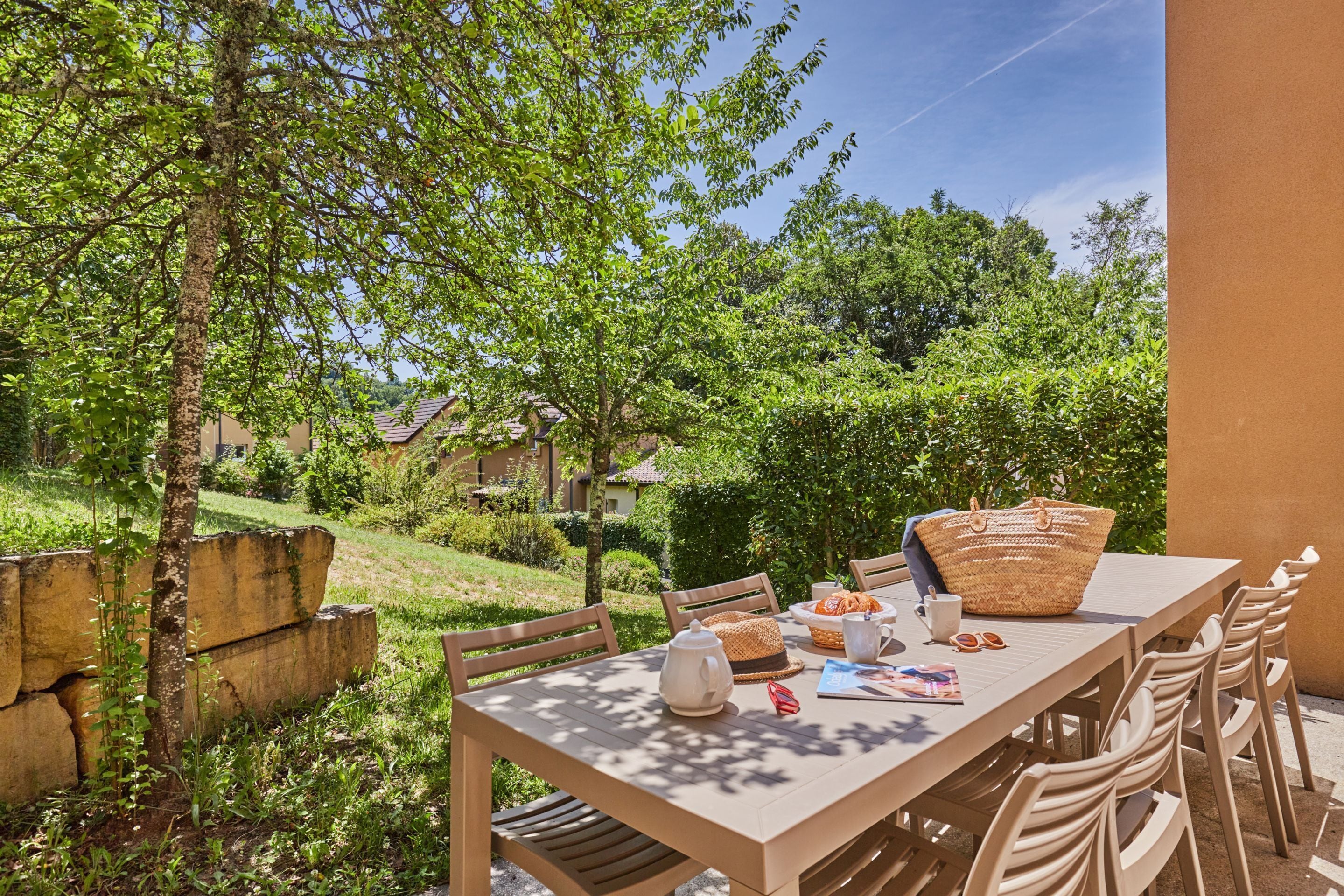 An outdoor dining area with a table set for two, surrounded by chairs, with a view of a garden and buildings in the background.