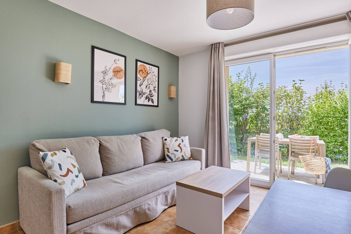 A living room with a sofa, a coffee table, and a dining area with chairs and a table. There are framed pictures on the wall and a window with curtains.