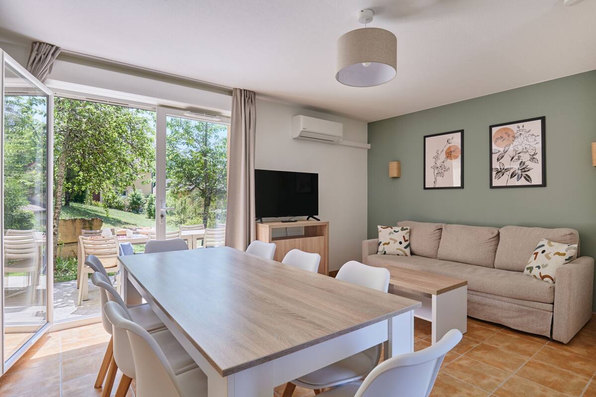 A modern living room with a wooden dining table, white chairs, a beige sofa, and a flat-screen TV.