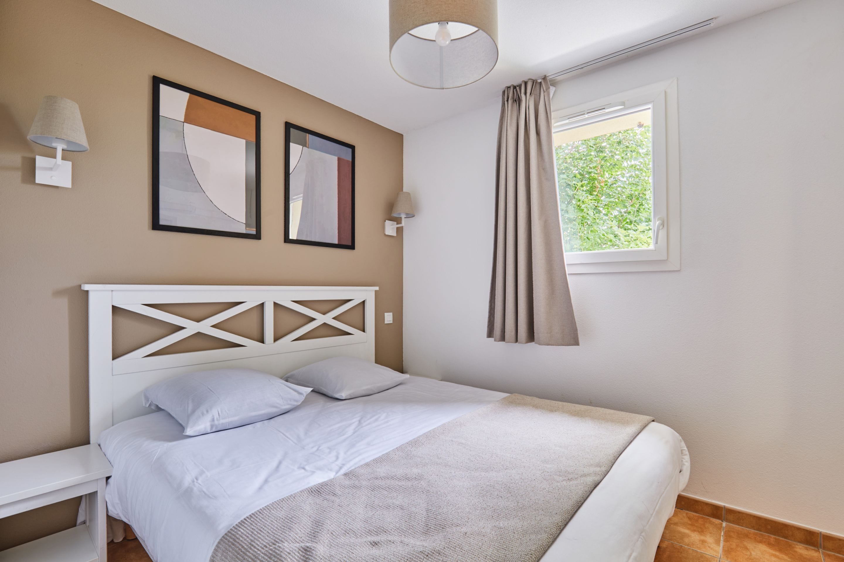 Une chambre à coucher comprenant un lit, une table de chevet, une fenêtre avec des rideaux et deux lampes fixées au mur.