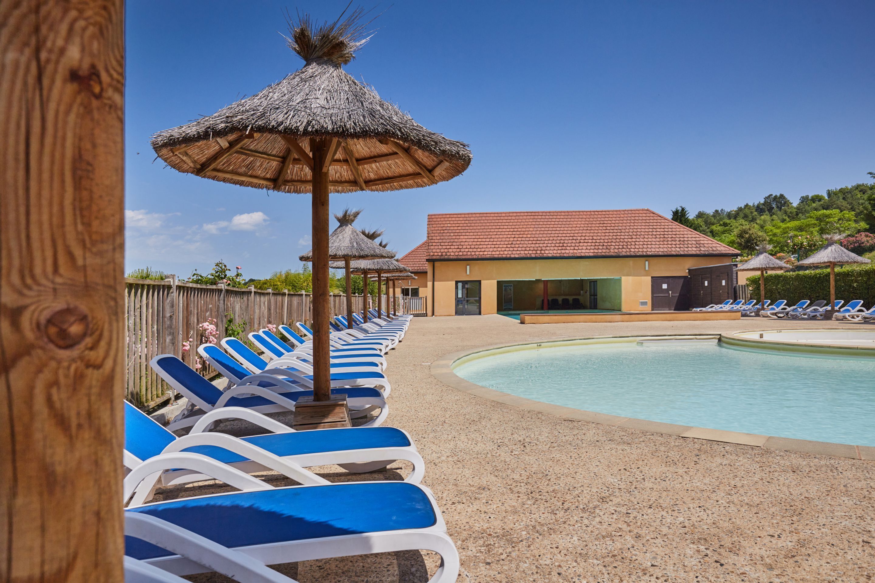 A pool area with lounge chairs and thatched umbrellas, a building with a red roof, and a wooden fence.