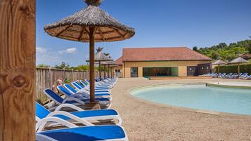 A pool area with lounge chairs and thatched umbrellas, a building with a red roof, and a wooden fence.