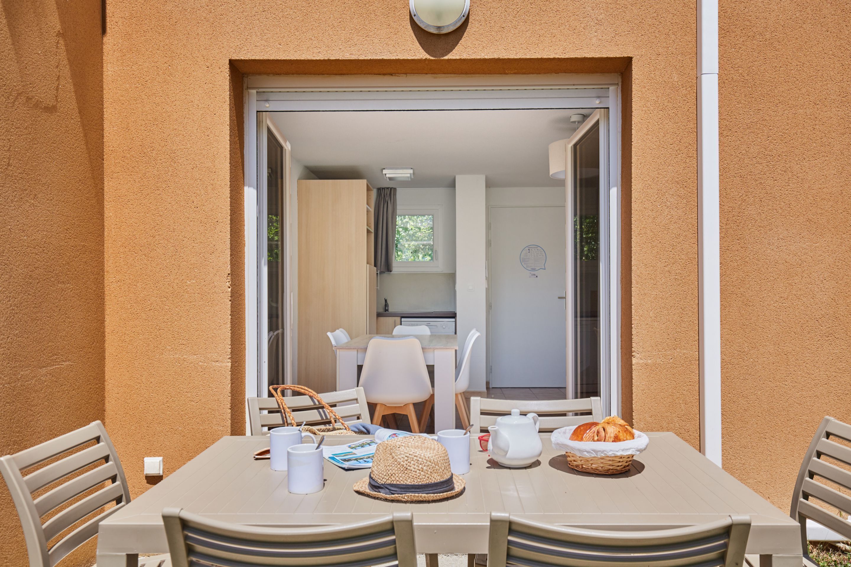 Un coin repas avec une table dressée pour deux personnes, des chaises, un panier de pain et un chapeau posé sur la table.