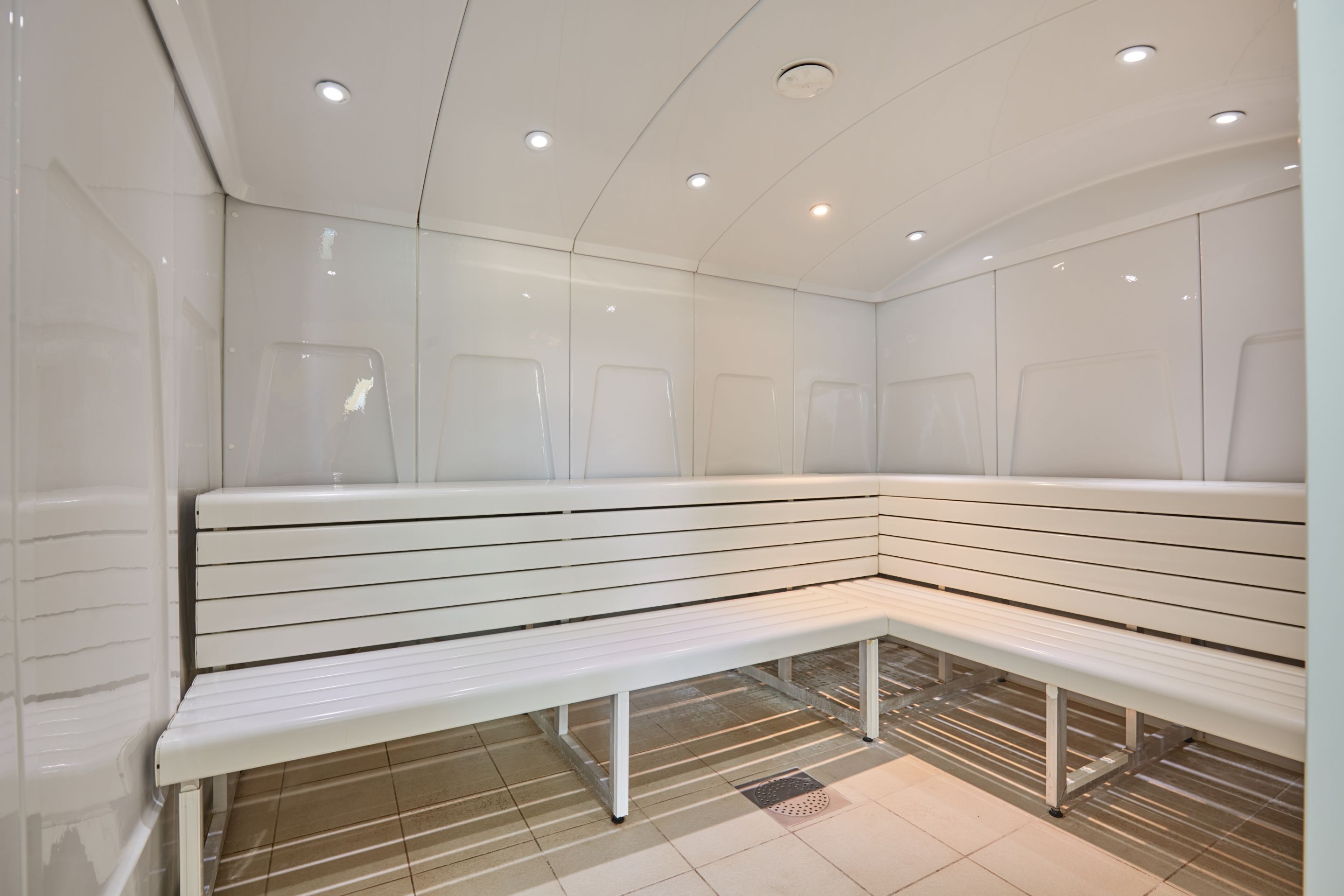 Un sauna moderno y limpio con un banco blanco y otros bancos, luces empotradas en el techo y suelo de baldosas.