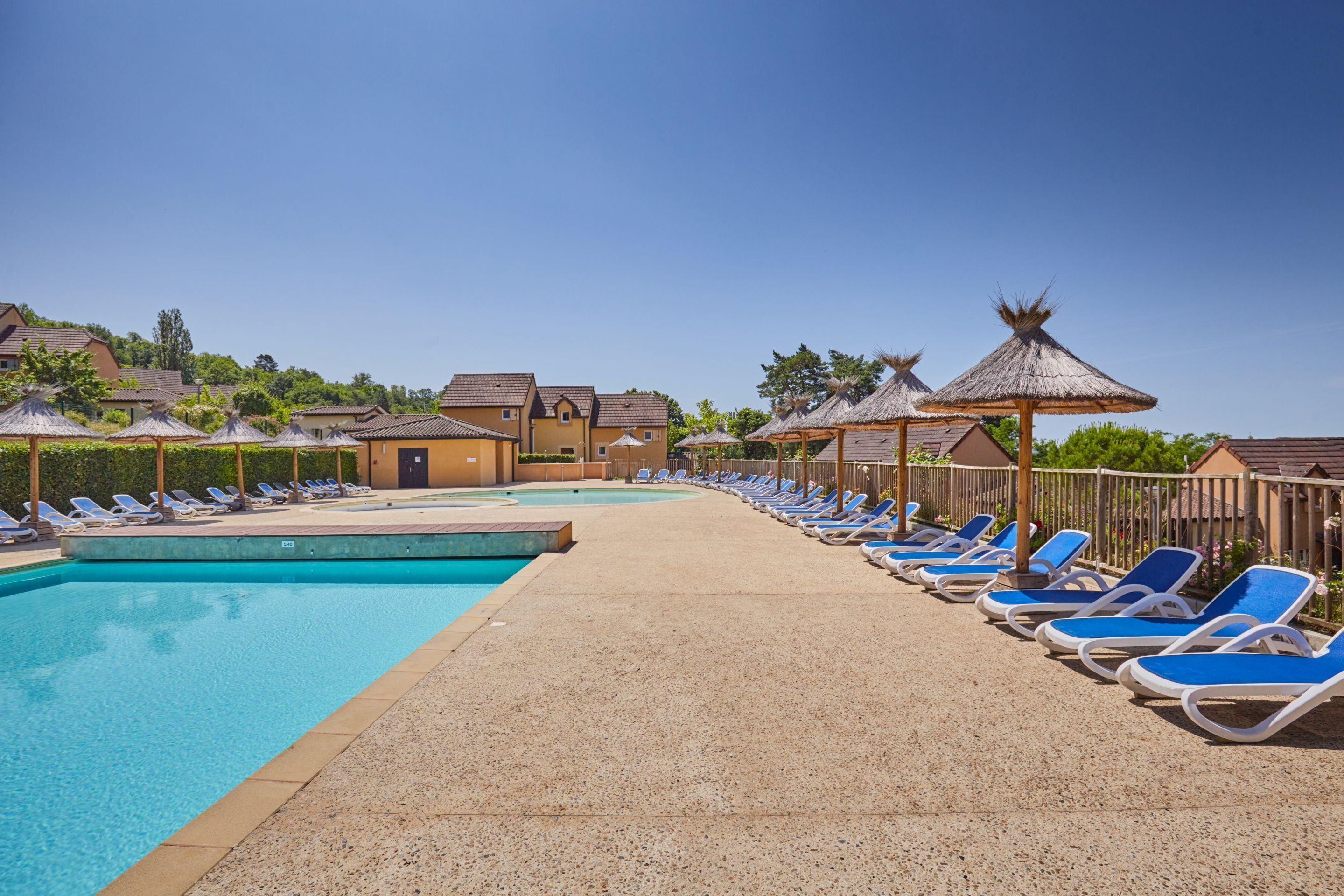 Une piscine entourée de bâtiments au style architectural uniforme, avec des chaises longues bleues et des parasols en chaume.