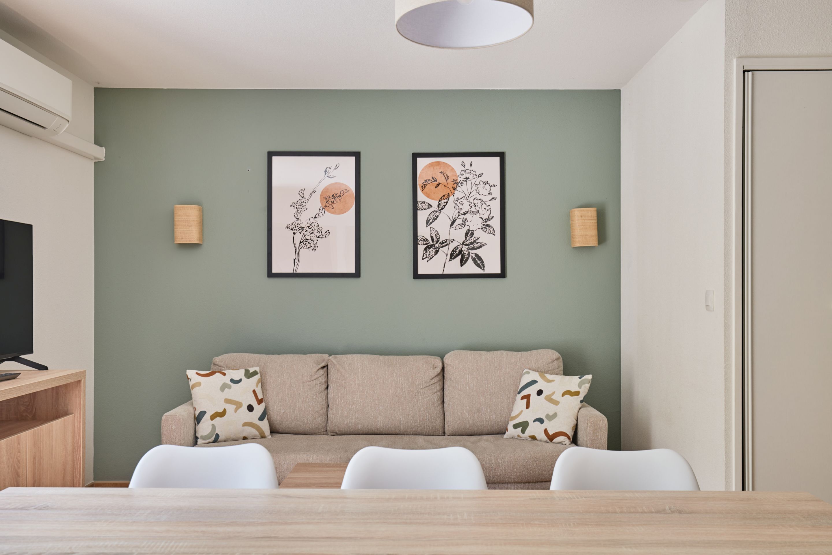 Una sala de estar moderna con un sofá, dos cuadros enmarcados con motivos florales, un televisor y una zona de comedor con una mesa y sillas de madera.