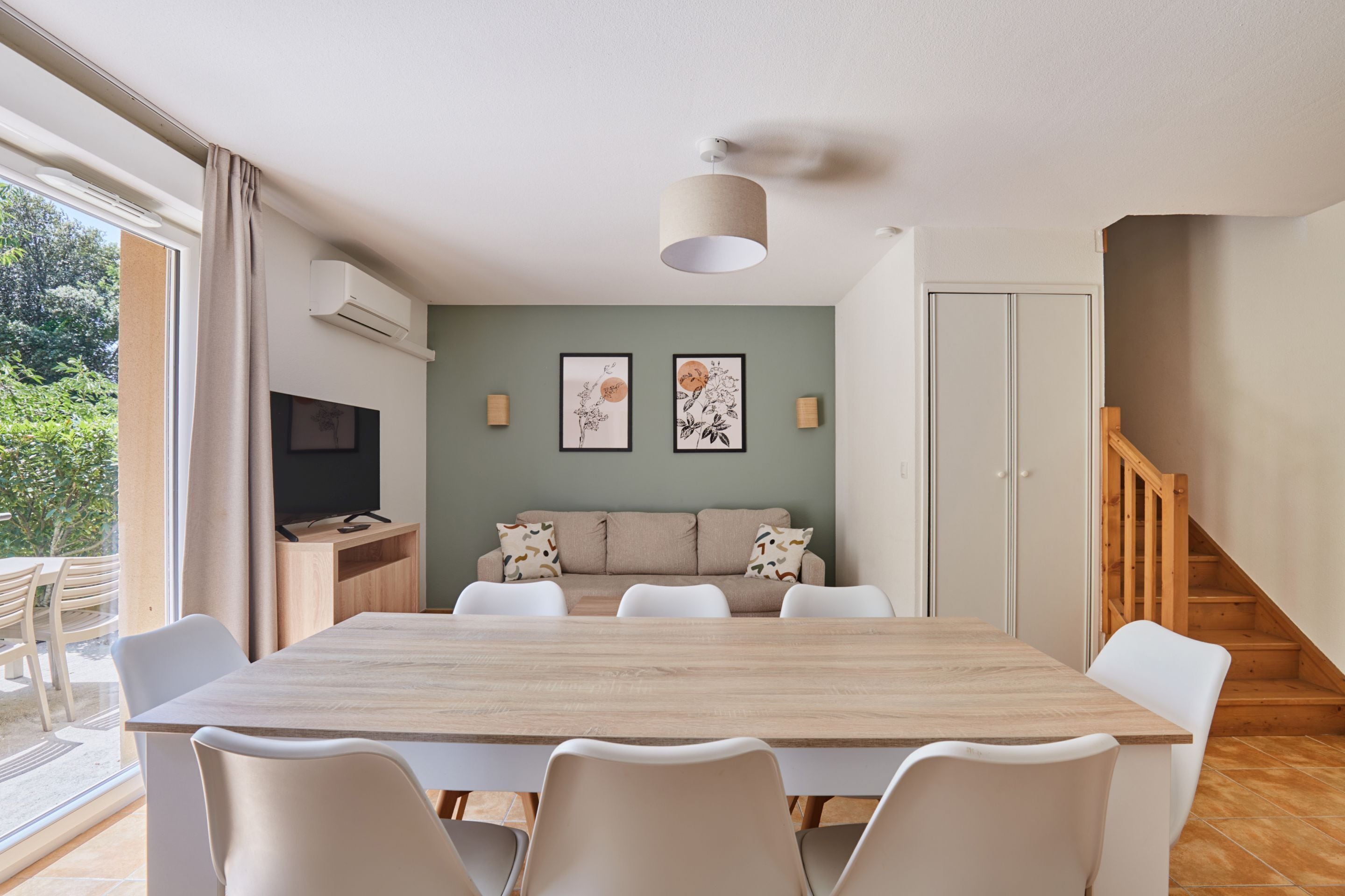 Un comedor con mesa de madera y sillas blancas, un sofá con cojines estampados y un televisor sobre un mueble de madera.
