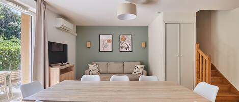 Un comedor con mesa de madera y sillas blancas, un sofá con cojines estampados y un televisor sobre un mueble de madera.