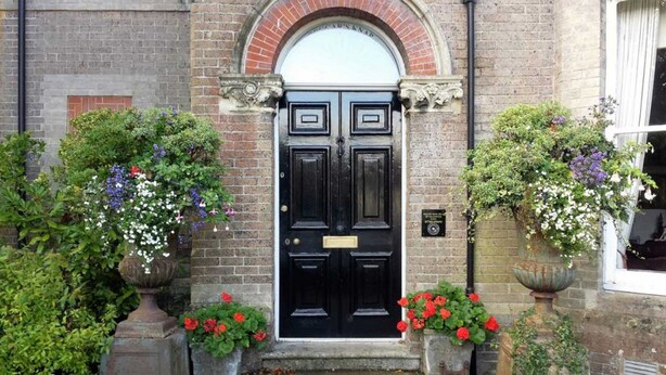 A black double door with a brick archway above it, flanked by two flower boxes with red and purple flowers. - Beggar's Knap (Dorchester)