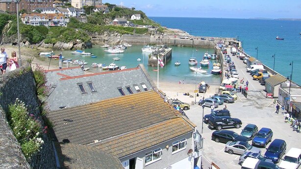 Un pueblo costero con un puerto deportivo, coches aparcados y edificios a lo largo del paseo marítimo.