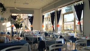A restaurant with multiple tables and chairs, large windows with curtains, and a ceiling fan.