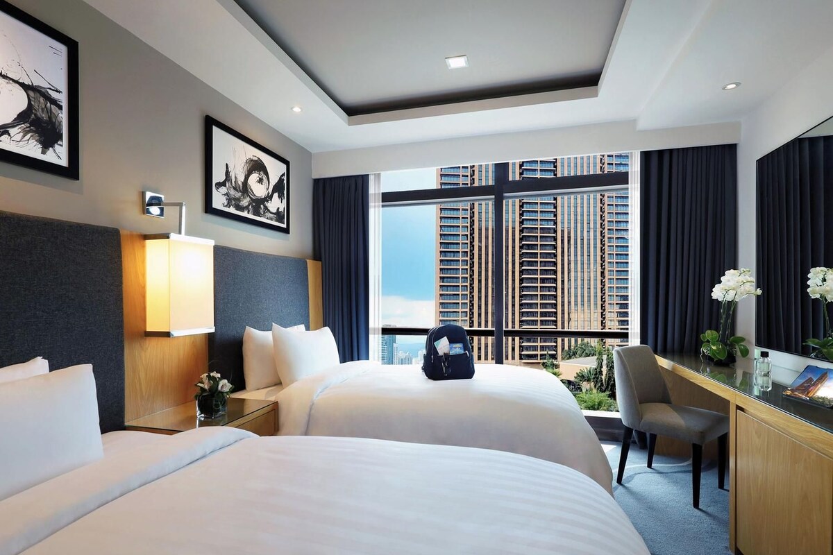 Een hotelkamer met twee bedden, een bureau, een stoel, een groot raam met uitzicht op de stad en kunstwerken aan de muur.
