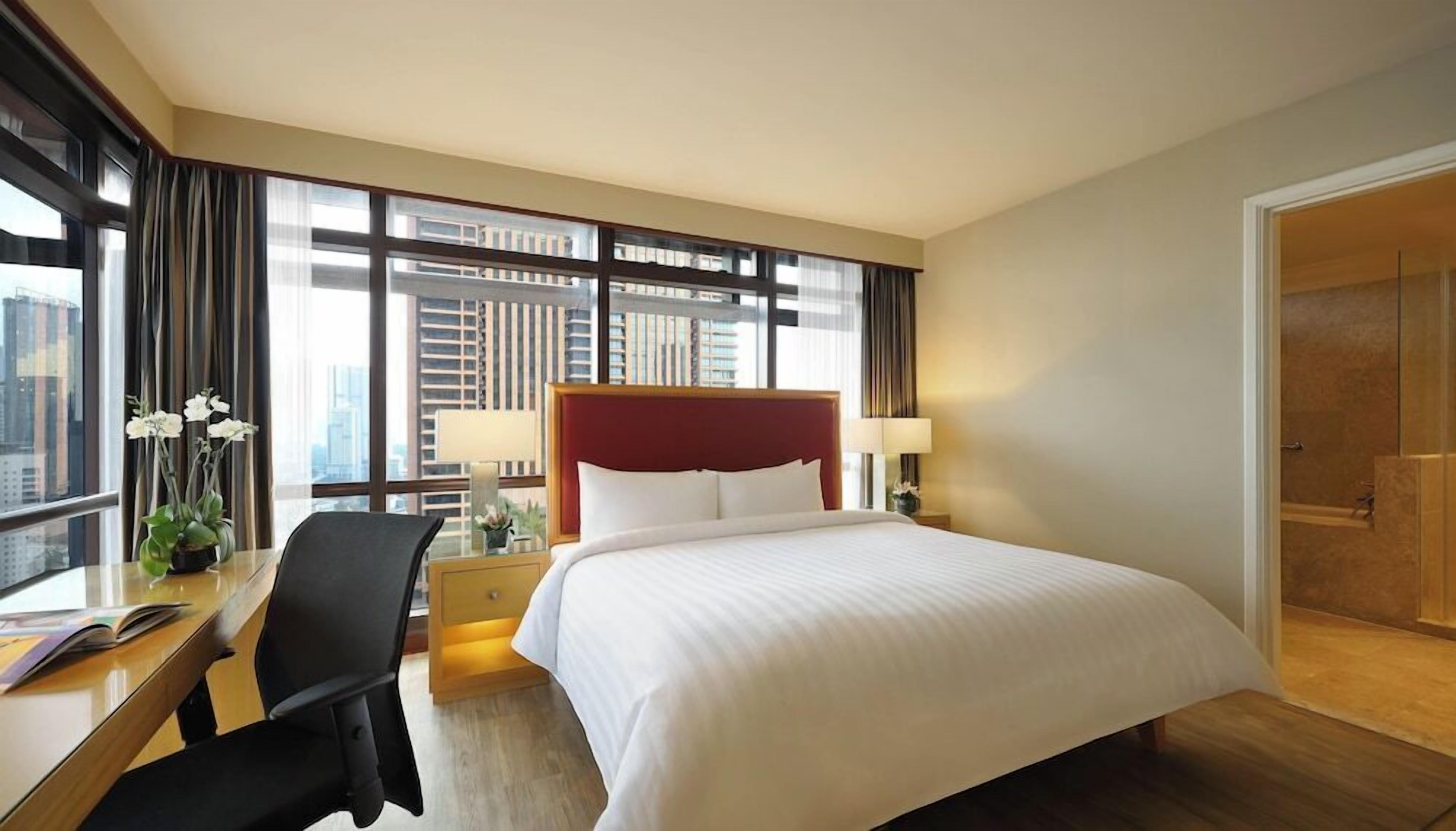 A hotel room with a large bed, a desk with a chair, two bedside lamps, a window with curtains, and a view of buildings.