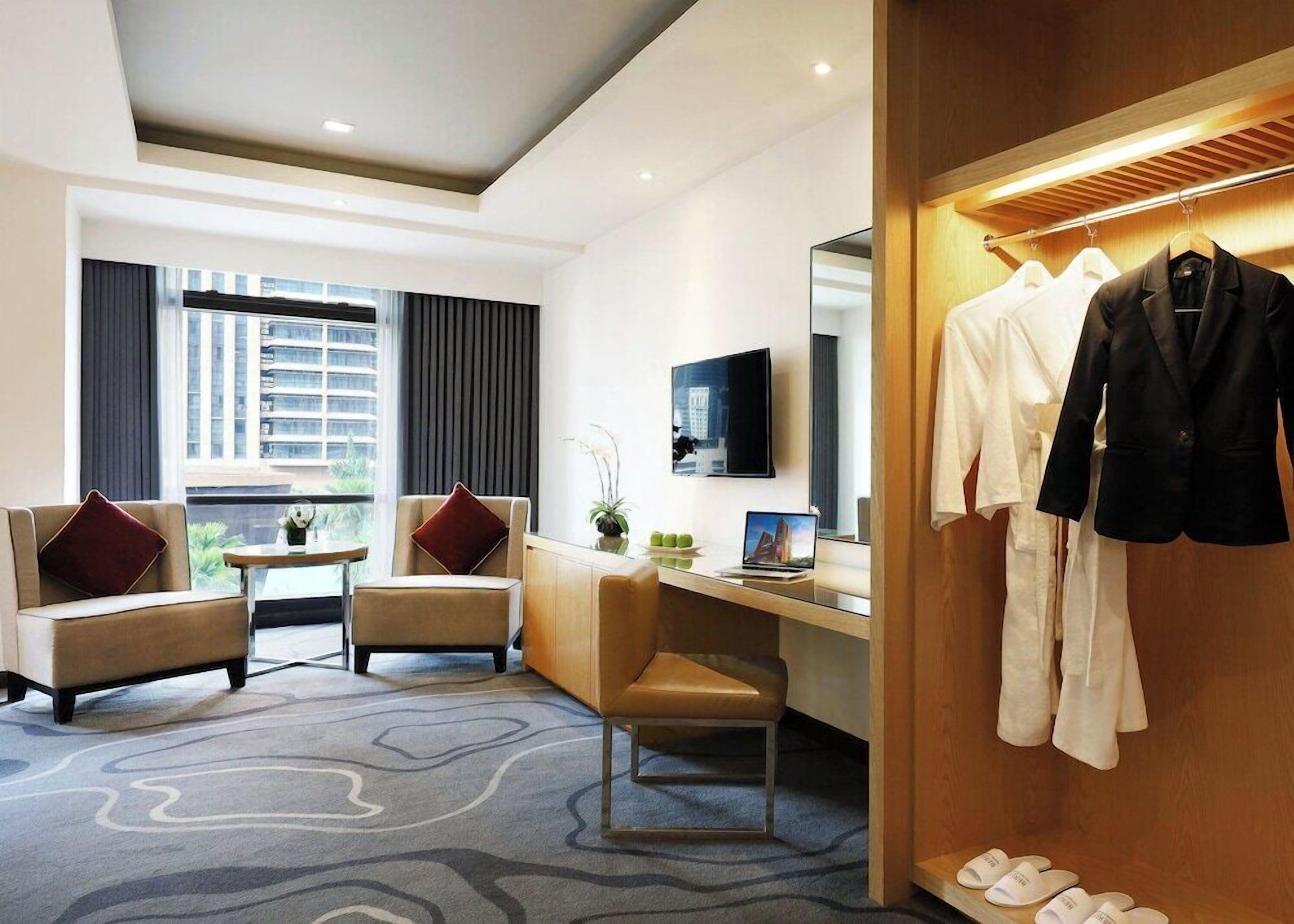 Une chambre d’hôtel équipée d’un canapé, d’un fauteuil, d’un bureau, d’un ordinateur portable, d’une télévision et d’un portant à vêtements avec des peignoirs et un costume.