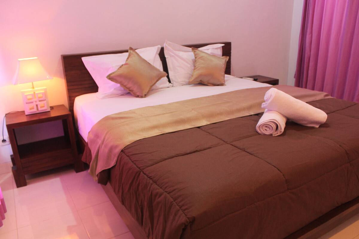 A neatly made bed with a brown bedspread and pillows, a small bedside table with a lamp, and a rolled towel on the bed.