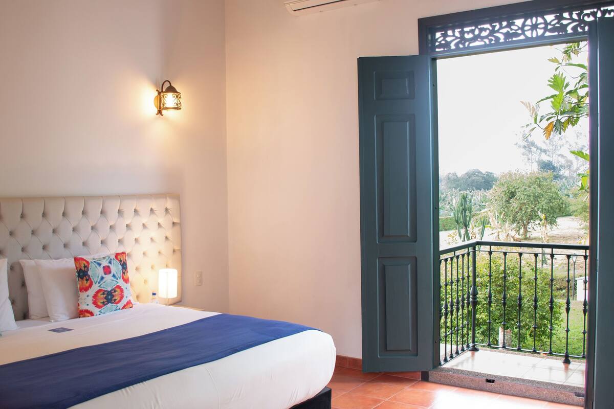 A bedroom with a tufted headboard, a bed with white and blue bedding, a colorful pillow, a wall-mounted light, and a balcony with a view of greenery.
