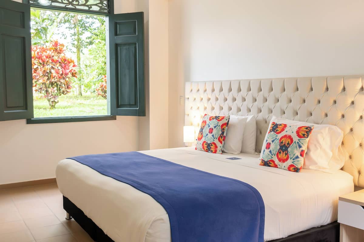 A neatly made bed with a blue bedspread, two patterned pillows, and a tufted headboard. A window with green shutters is visible, showing a garden outside.
