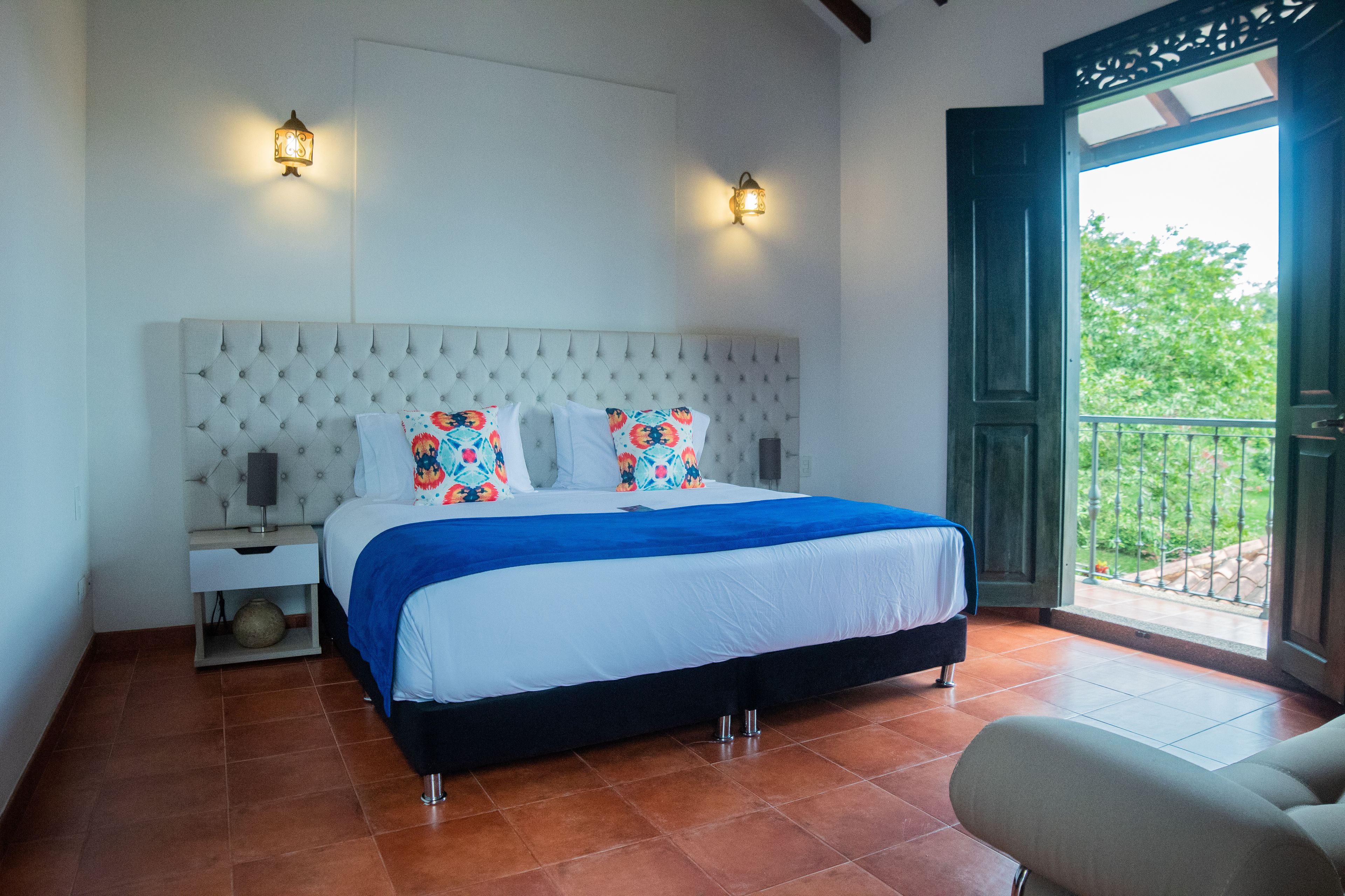 A bedroom with a tufted headboard, a bed with a blue blanket, two wall-mounted lamps, and a view of greenery through an open door.