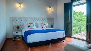 A bedroom with a tufted headboard, a bed with a blue blanket, two wall-mounted lamps, and a view of greenery through an open door.