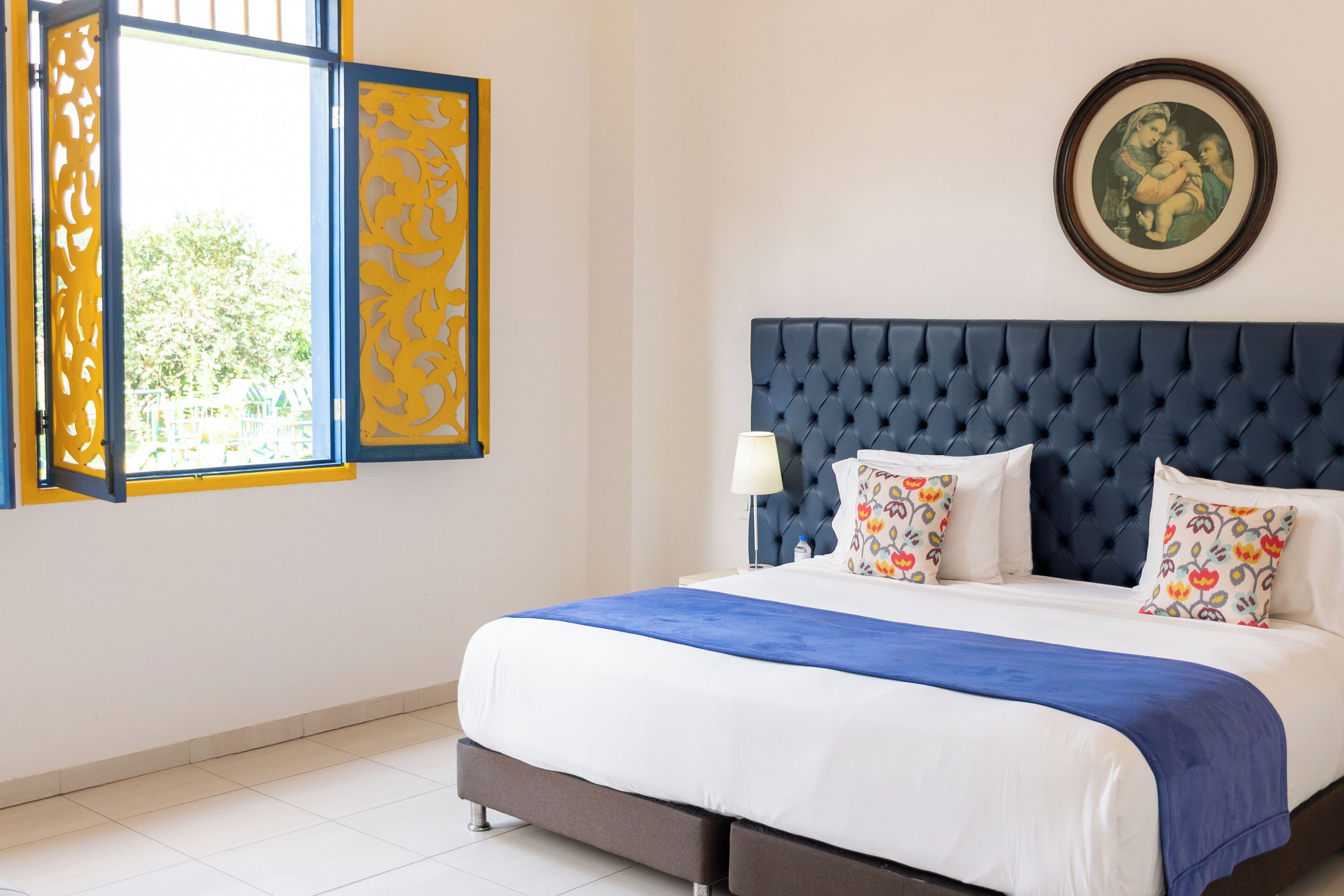 A neatly made bed with a blue headboard, a decorative window with yellow and blue patterns, and a framed picture on the wall.