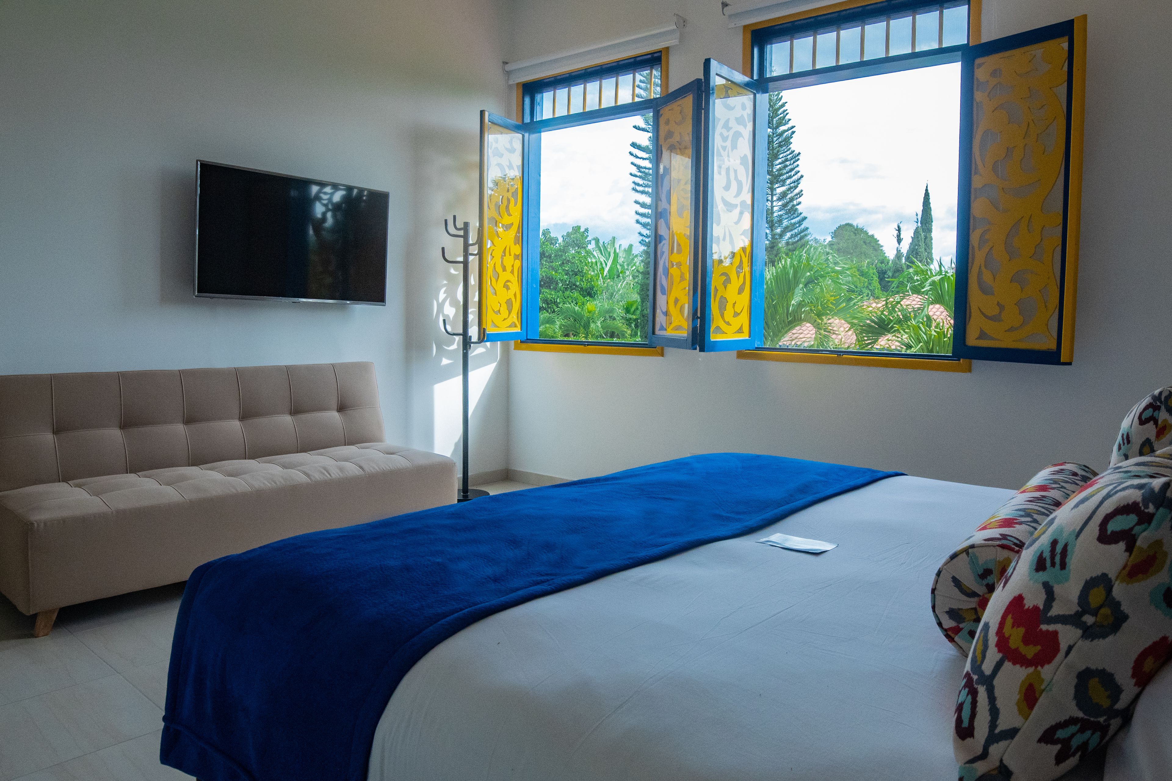 A bedroom with a bed, a sofa, a TV, and a window with a view of greenery.