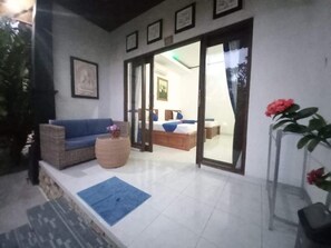 Una habitación con sofá, cama y una pequeña mesa con un florero. Se observan cuadros en la pared y una planta a la derecha.