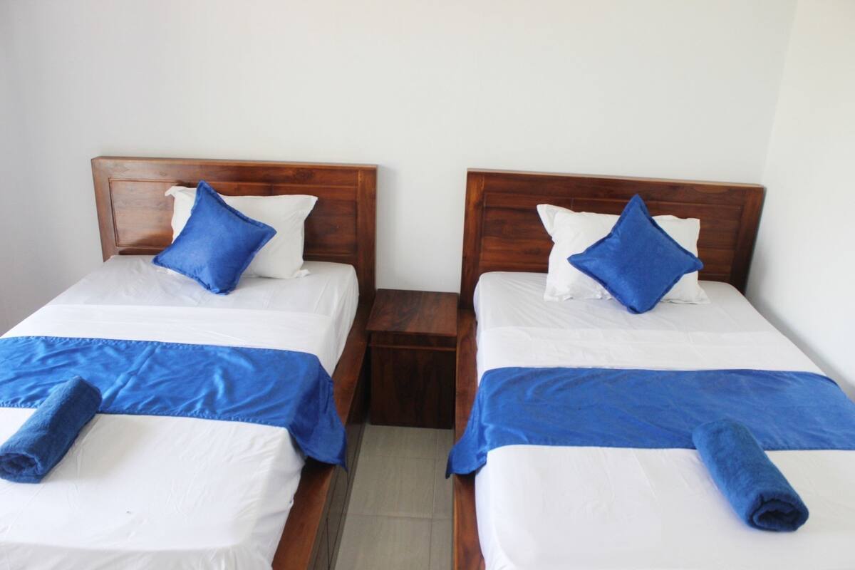 Duas camas de solteiro com cabeceiras de madeira, roupa de cama branca e detalhes em azul.