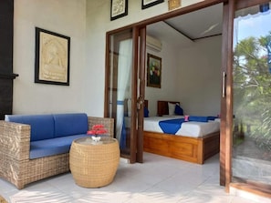 A room with a sofa, a round wicker table, and a bed with a blue and white bedspread. - Bobo Bungalow (Lembongan Island)