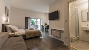A modern hotel room with a bed, a sofa, a TV, a bathroom with a shower, and a view of the outside. - King's Street Apartments (Zagreb)