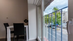 A room with a dining table and chairs, a potted plant, and a large window overlooking a street with parked cars and buildings. - King's Street Apartments (Zagreb)