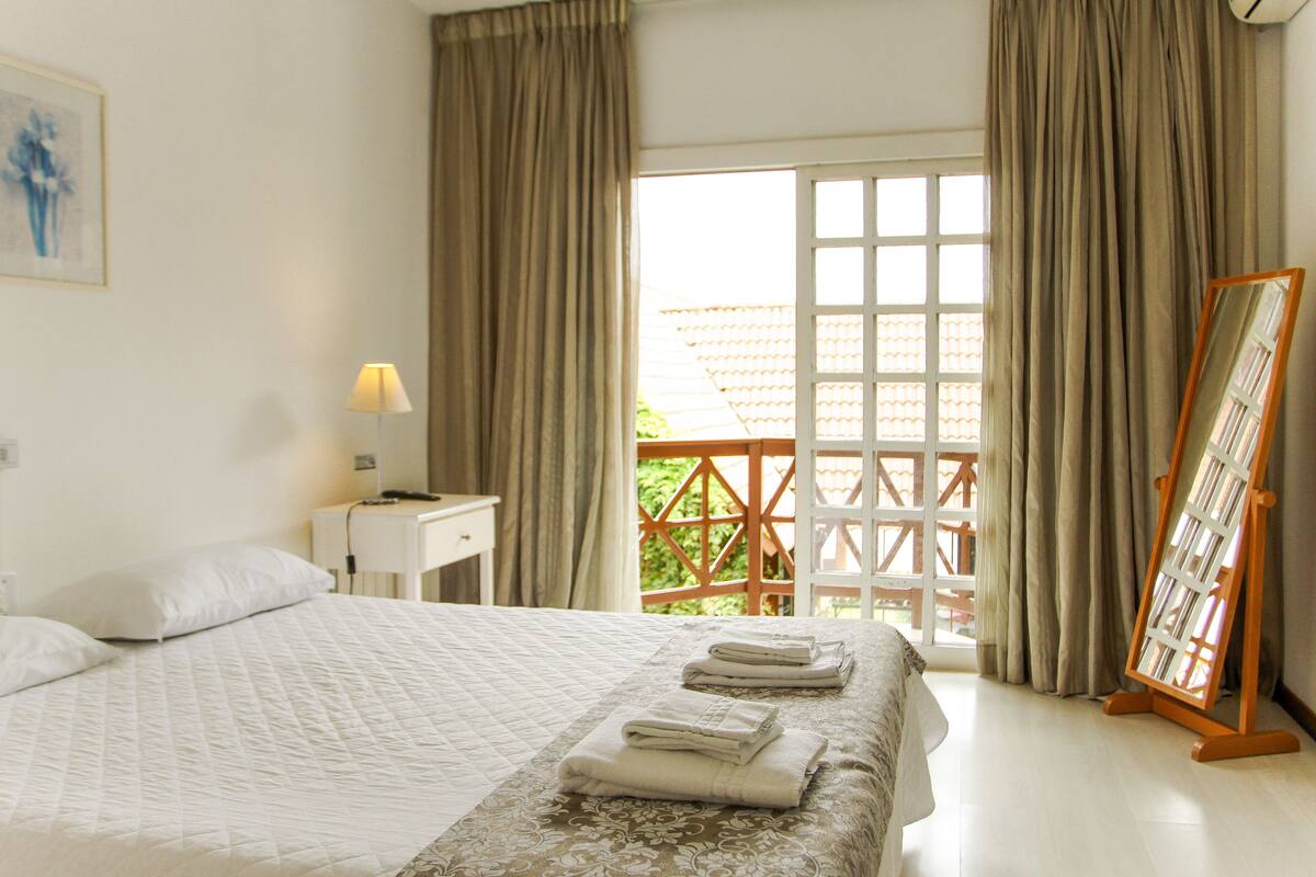 Una cama bien hecha con cubrecama acolchado, una mesita de noche con lámpara y una ventana con cortinas.