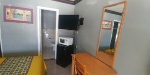 Um quarto pequeno com uma cama, uma mesa de madeira, um espelho, uma TV e um micro-ondas.