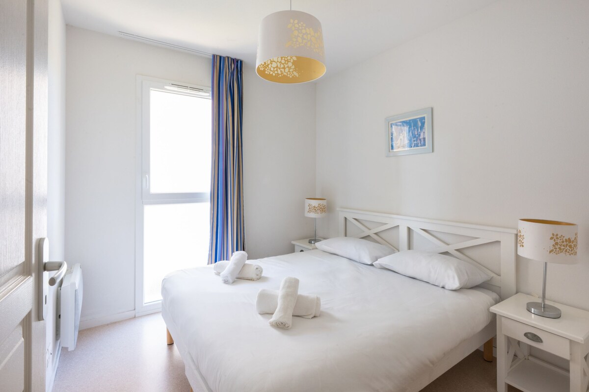 Habitación de hotel con una cama grande, dos mesitas de noche, una lámpara y una ventana con cortinas.
