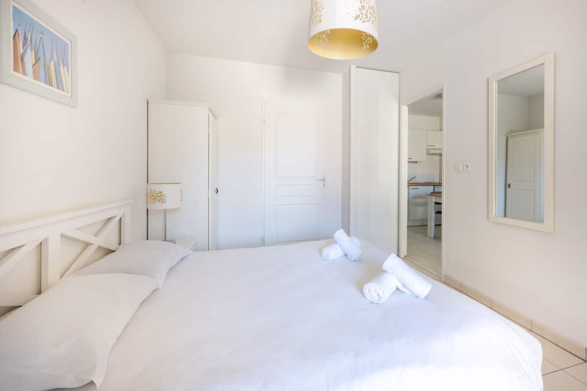 A bedroom with a bed, a door, a mirror, and a hallway leading to a kitchen.