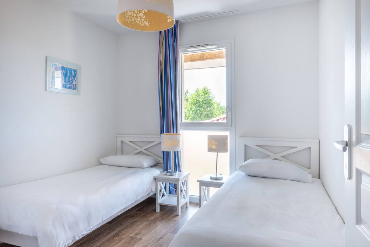 A hotel room with two single beds, a small table, a lamp, a window with curtains, and a picture on the wall.
