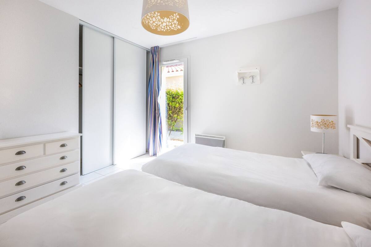 A modern bedroom with two beds, a white dresser, a ceiling light fixture, and a view of the outdoors.