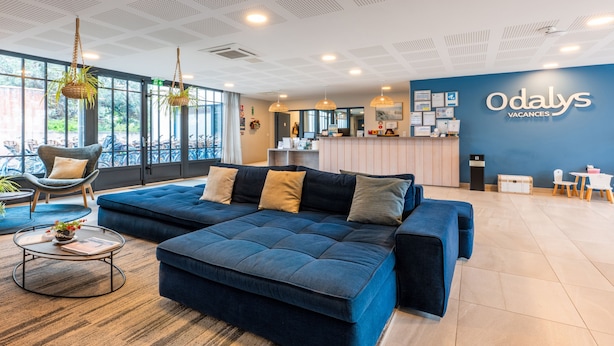 A modern hotel lobby with a large blue sofa, a round coffee table, and a reception desk. - Résidence Odalys Rêve d'lle (Rivedoux-Plage)