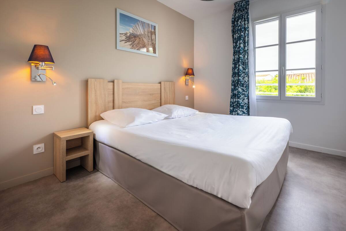 Ein ordentlich bezogenes Bett mit Holz Kopfteil, zwei Nachttischlampen, ein kleiner Nachttisch und ein Fenster mit Blick auf Grünflächen.