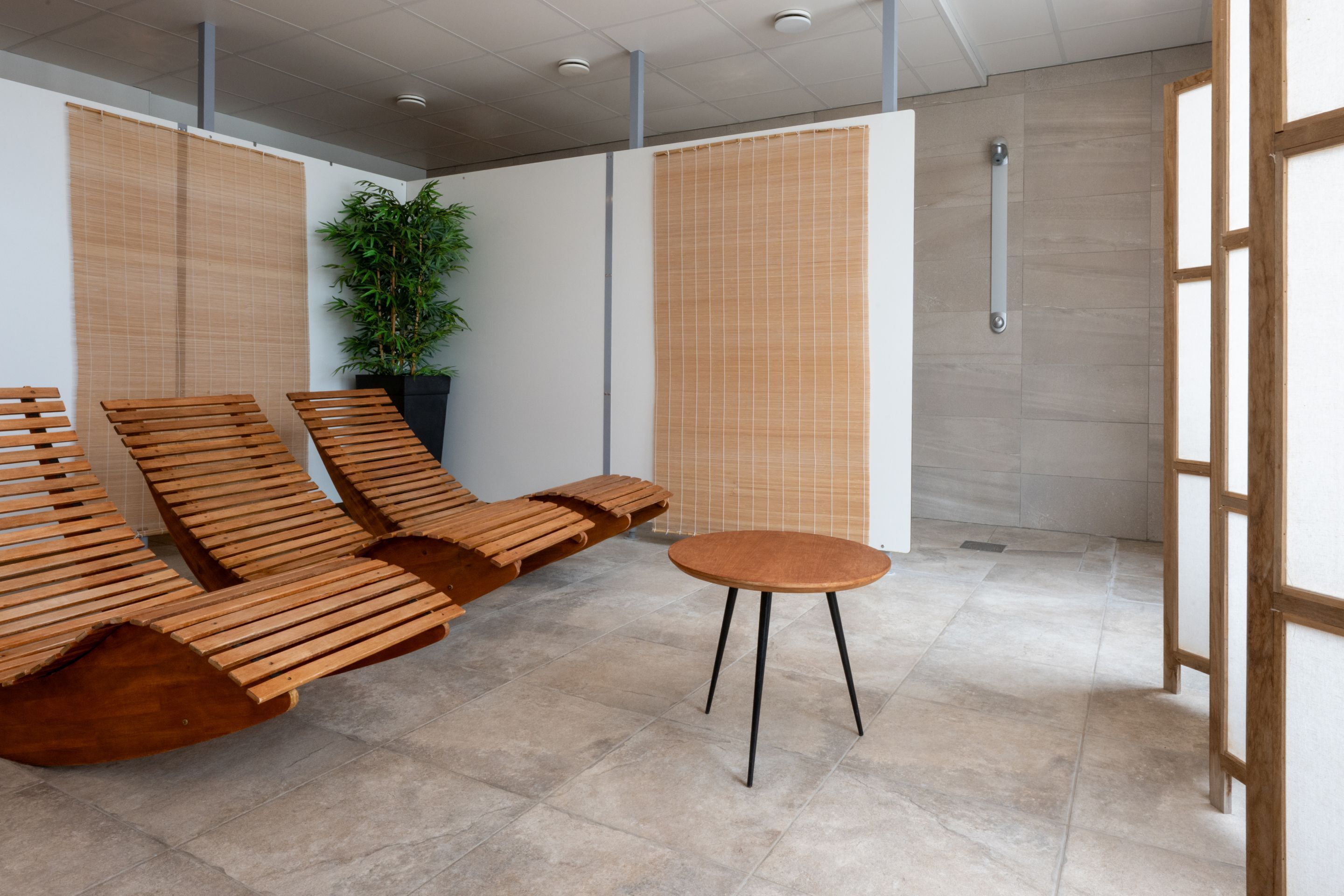 A modern waiting area with wooden lounge chairs and a small table, a potted plant, and a bamboo screen.