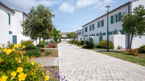 A paved pathway between white buildings with green shutters, surrounded by flowering plants and trees. - Résidence Odalys Rêve d'lle (Rivedoux-Plage)