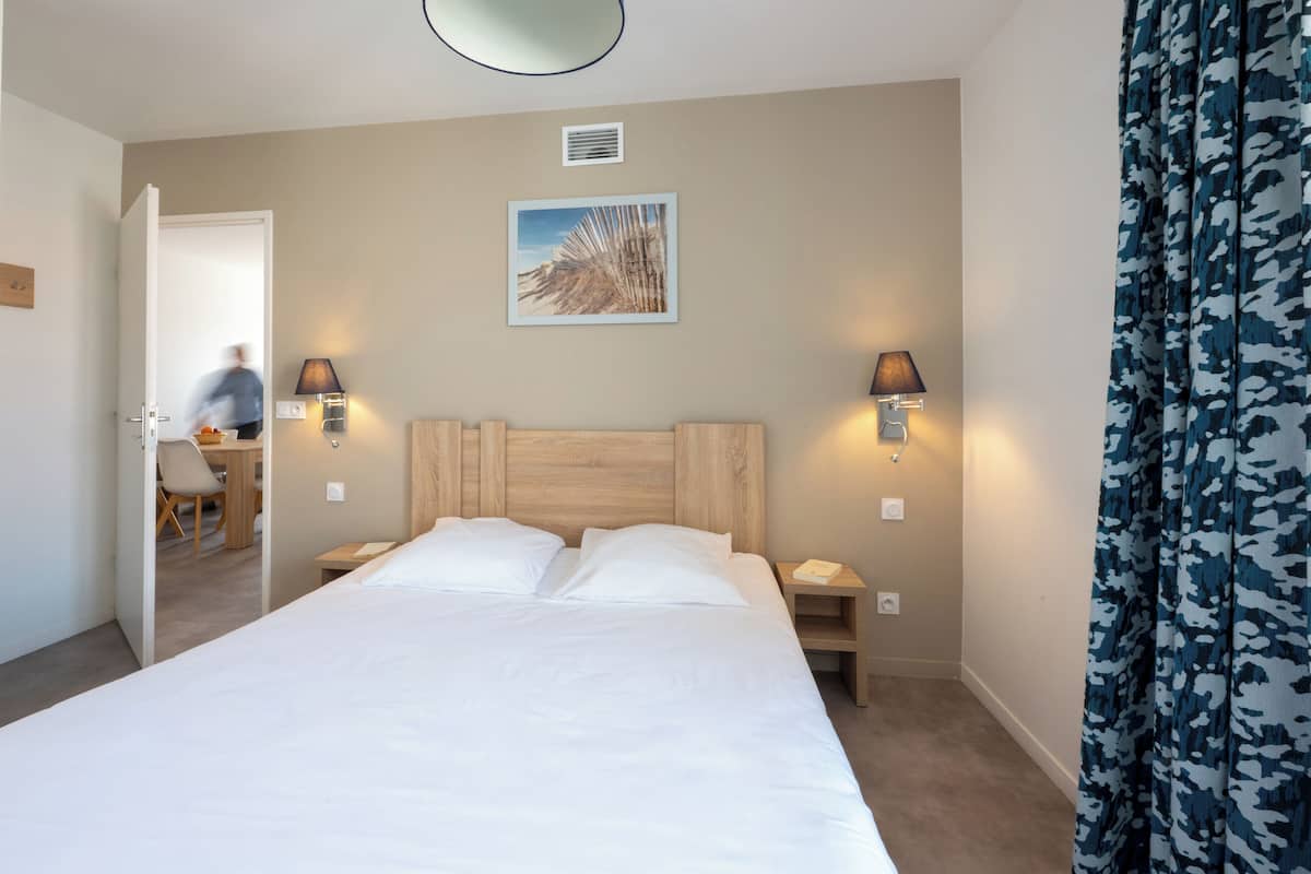 Ein Hotelzimmer mit einem großen Bett, zwei Nachttischen, einem Schreibtisch mit Stuhl, einem Wandbild und einer Tür zu einem weiteren Raum.
