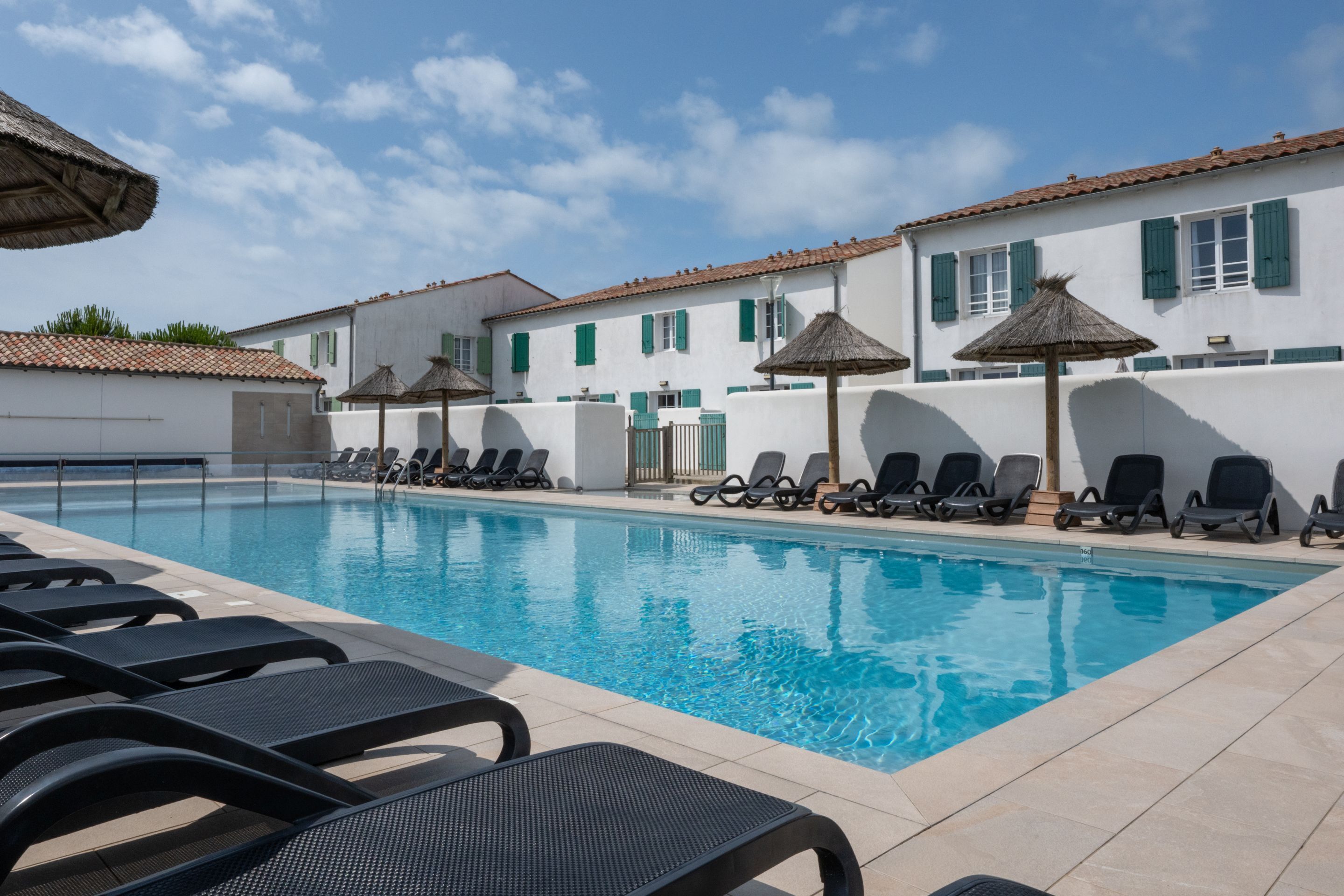 Una piscina con sillas de descanso y sombrillas, rodeada de edificios blancos con contraventanas verdes.