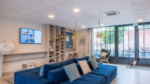 A modern living room with a blue sofa, a bookshelf, and a large window with a view of bicycles. - Résidence Odalys Rêve d'lle (Rivedoux-Plage)