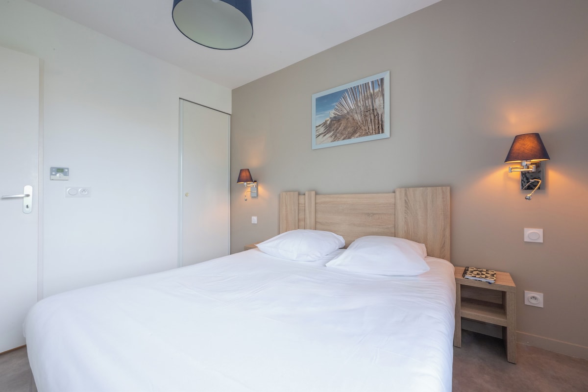 Una cama bien tendida con sábanas blancas en una habitación de hotel. Hay dos lámparas fijadas a la pared a cada lado de la cama.