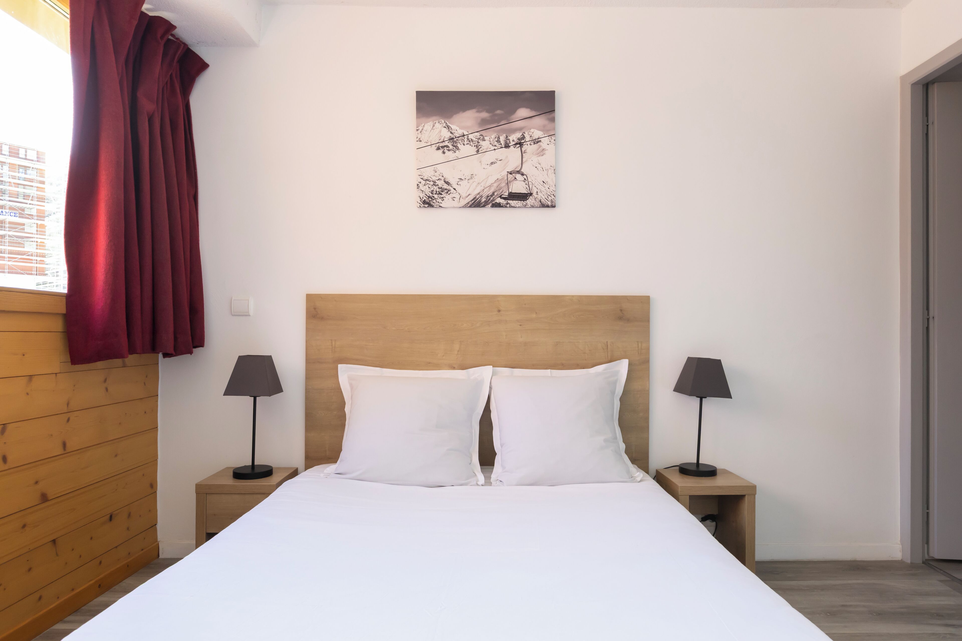 Un dormitorio con cabecera de madera, ropa de cama blanca, dos lámparas de noche y una obra de arte con paisaje de montaña en la pared.