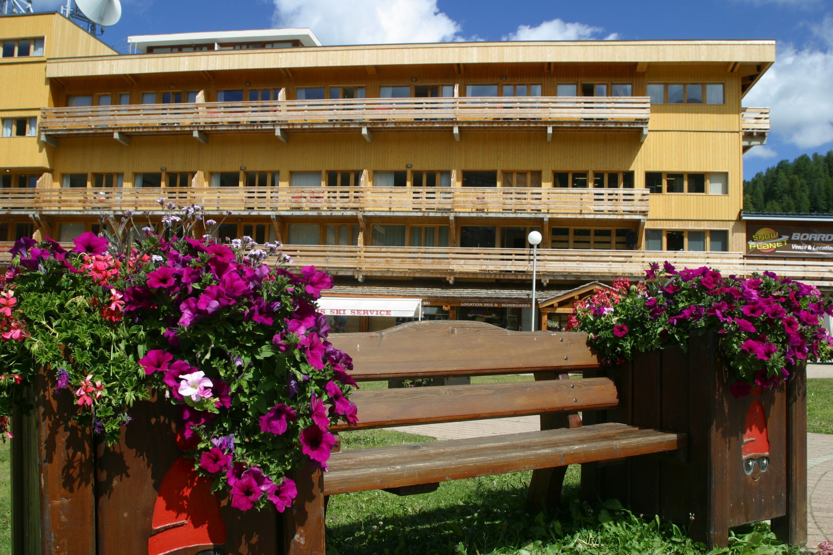 Un banco de madera con jardineras florales frente a un edificio de varios pisos con balcones.