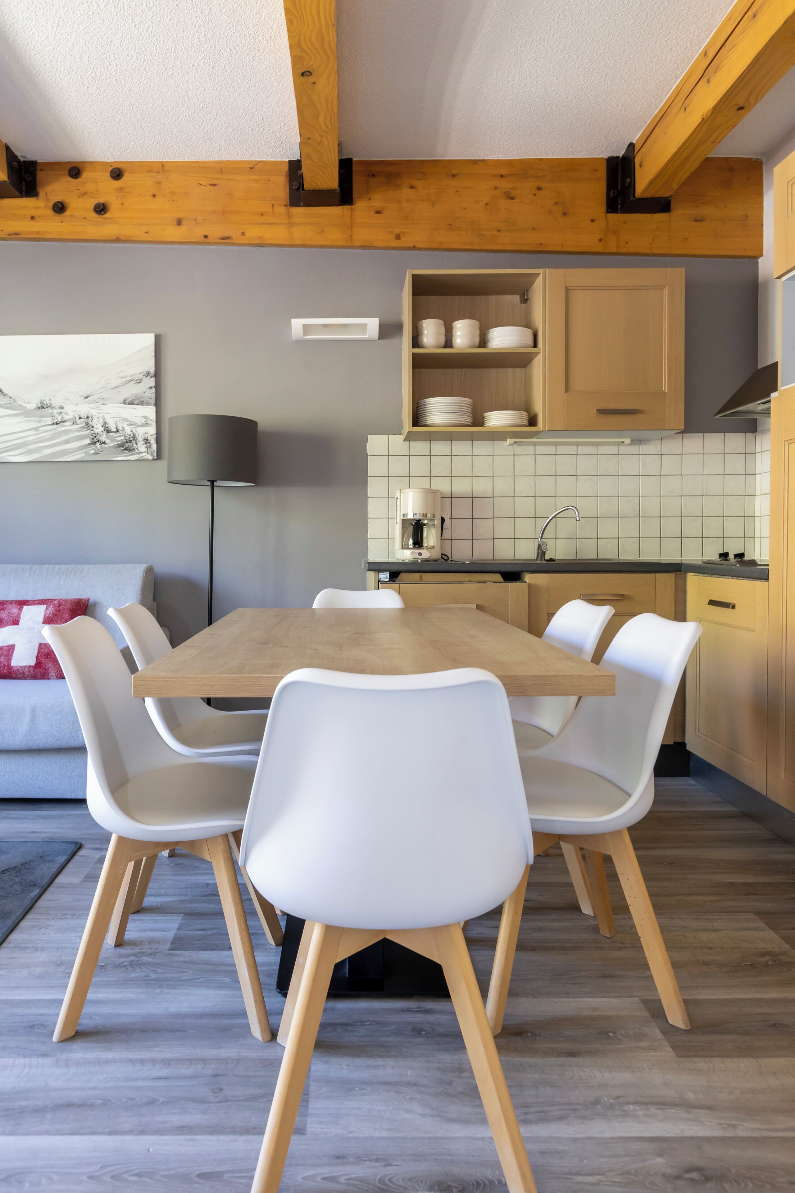 A dining area with a wooden table and white chairs, a kitchen with tiled backsplash, and a painting on the wall.