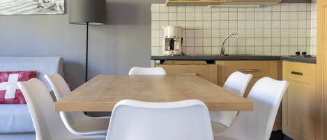 A dining area with a wooden table and white chairs, a kitchen with tiled backsplash, and a painting on the wall.